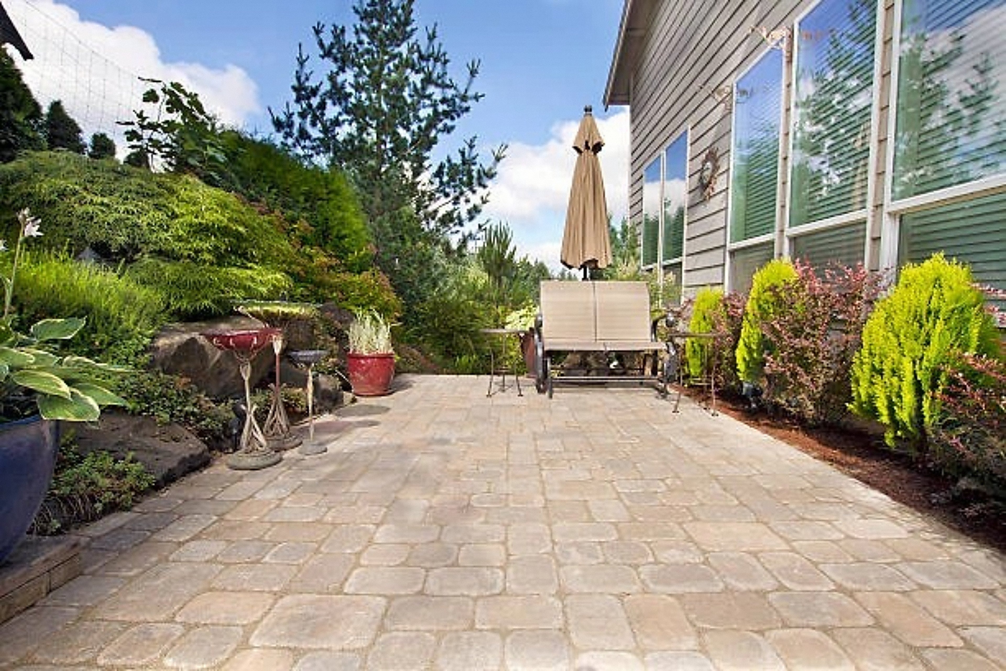 Paver patio with a bench and umbrella beside a garden bed and a home